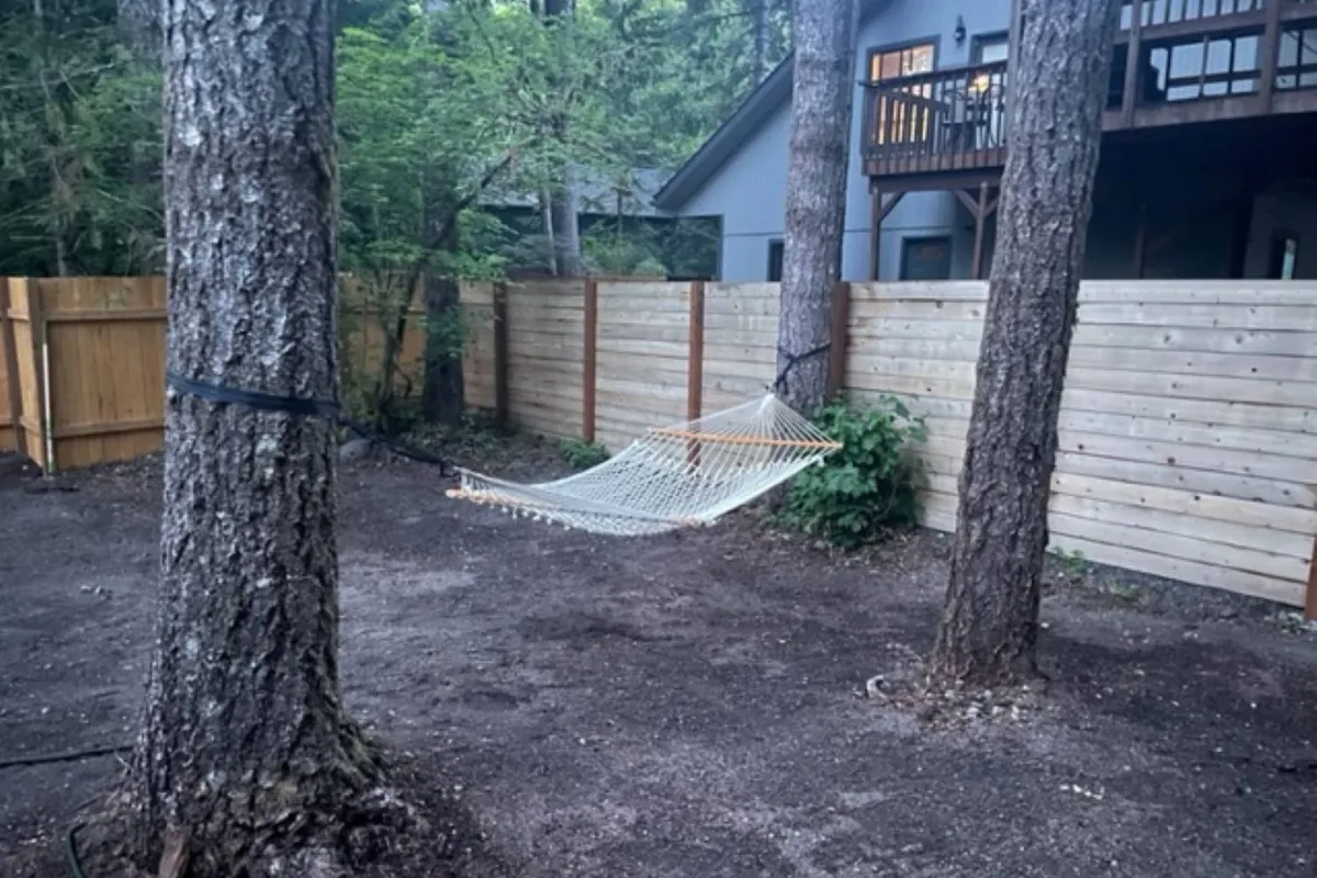 A peaceful outdoor scene with a hammock suspended between trees and a wooden fence enclosing the area. The fence is a prominent feature, and the tranquil environment showcases an ideal spot for pets and owners alike to enjoy nature.