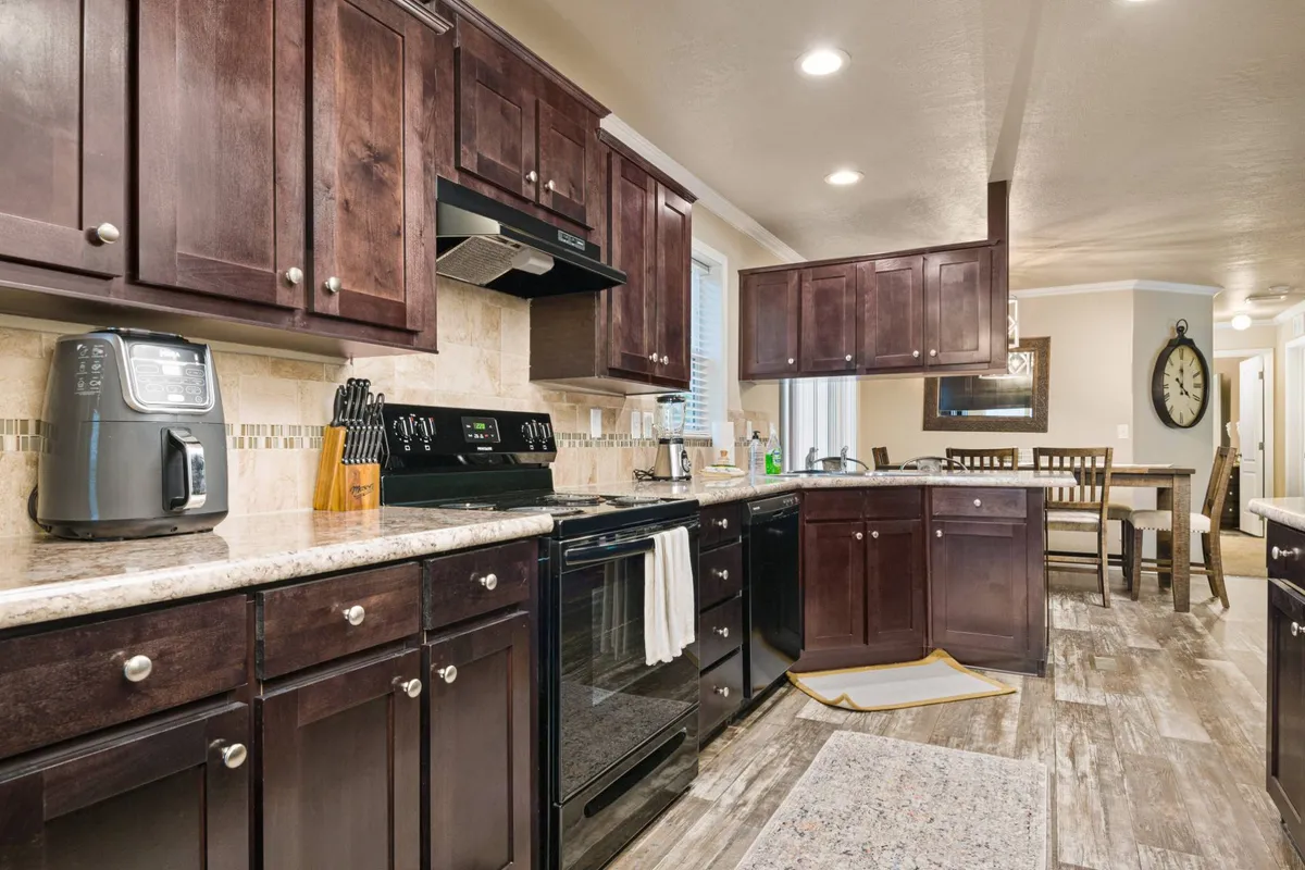 A warm, open-concept kitchen with dark wooden cabinets, a sleek granite countertop, and modern stainless steel appliances. The dining area is visible in the background, with ample space for gatherings, and the setup suggests a spot where cooking, socializing, and sharing meals seamlessly blend together.