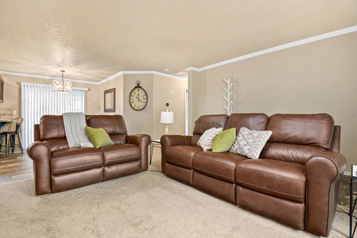 A bright and inviting living area featuring plush leather sofas adorned with decorative pillows, a stylish wall clock, a cozy lamp, and a verdant plant. The space is designed for relaxation after a long day spent outdoors, exuding a warm, welcoming vibe ideal for gathering with friends or family.