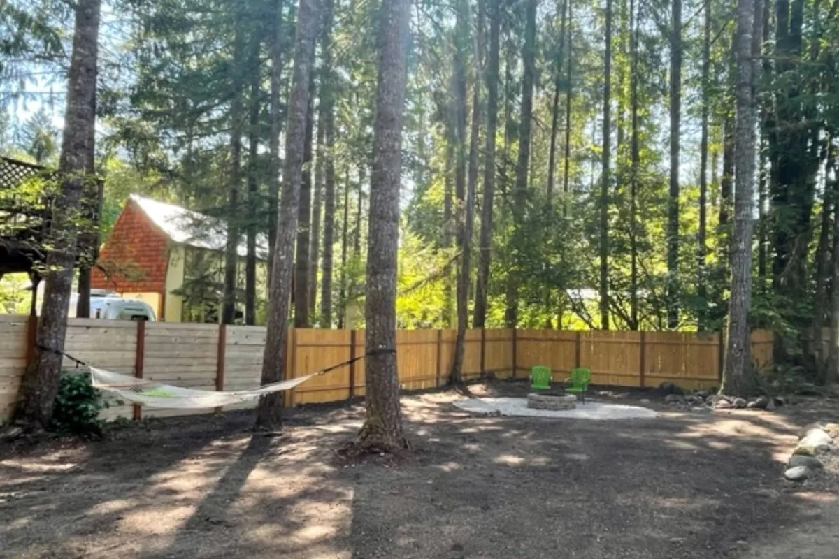 This image captures the backyard area featuring a wooden fence, tall trees, a hammock, a fire pit, and green chairs. The fenced perimeter is clearly visible, conveying both security and a natural setting ideal for pets. The arrangement invites relaxation and time spent outdoors.