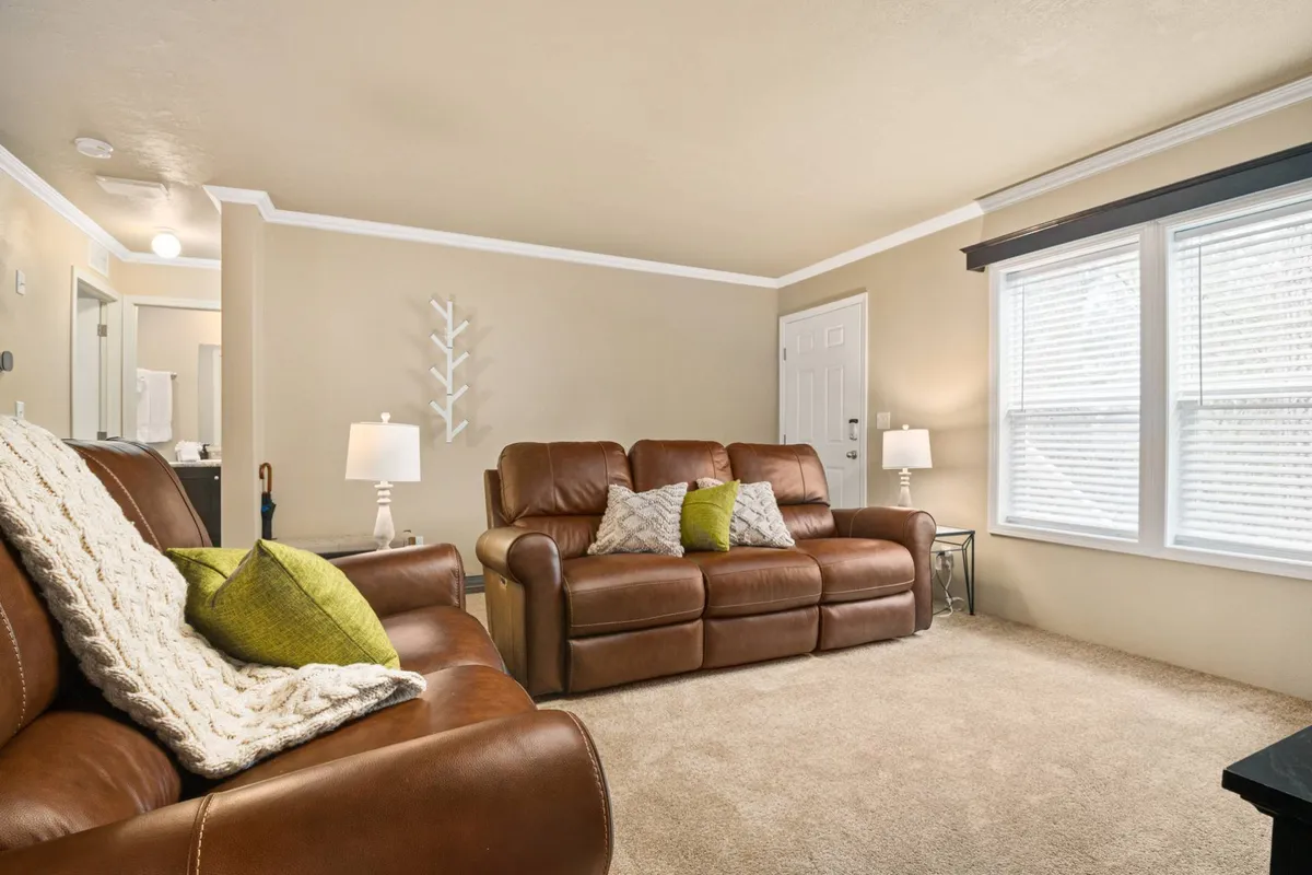 This image shows a cozy living room with leather sofas, decorative pillows, and soft lighting from table lamps. A window with blinds lets in gentle natural light, contributing to a relaxed atmosphere ideal for family gatherings or downtime after enjoying nearby amenities.