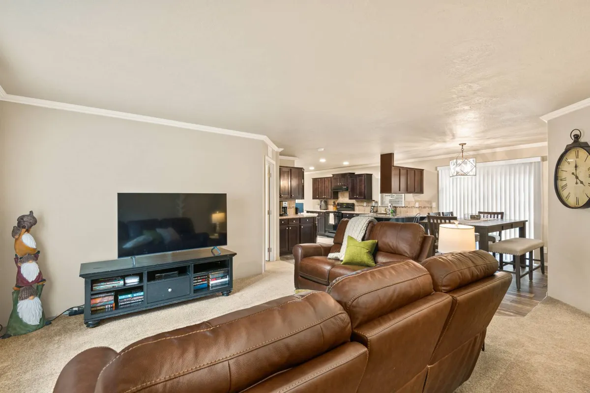 This image features a spacious, sunlit open-concept living area with a leather sofa facing a television, a dining table set for gatherings, and a modern kitchen visible in the background. Expansive windows allow natural light to pour in, highlighting the comfortable and contemporary furnishings.